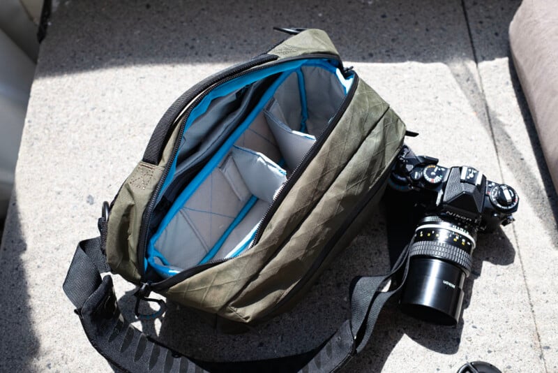 An olive green camera bag with a blue-lined, padded interior sits open on a concrete surface next to a black camera with a lens attached. The bag has adjustable dividers inside.