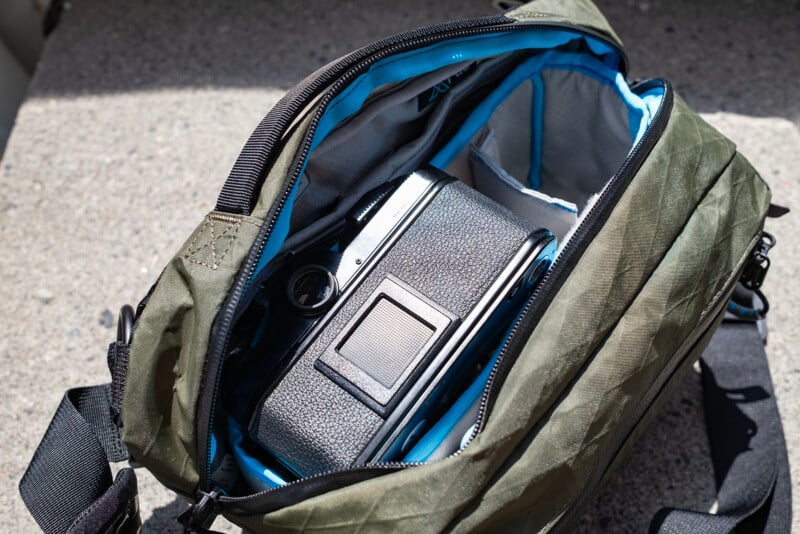 An open green camera bag with blue lining sits on a rough surface, revealing a black film camera stored inside the main compartment.