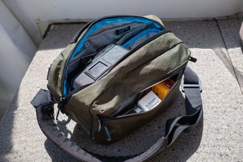 An olive green camera bag with a black strap is unzipped, revealing a camera and several boxes of film inside, resting on a concrete surface in bright sunlight.