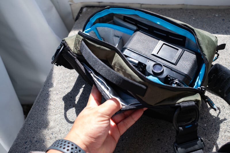 A hand opens a green camera bag, revealing a black camera and organized compartments inside. The bag has blue lining and sits on a gray surface in sunlight.