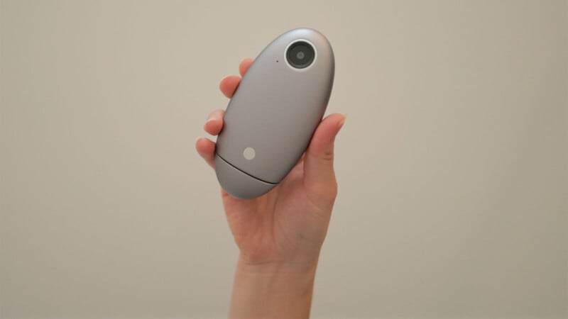 A hand holding a small, oval-shaped, silver electronic device with a circular lens in the upper corner against a plain light background.