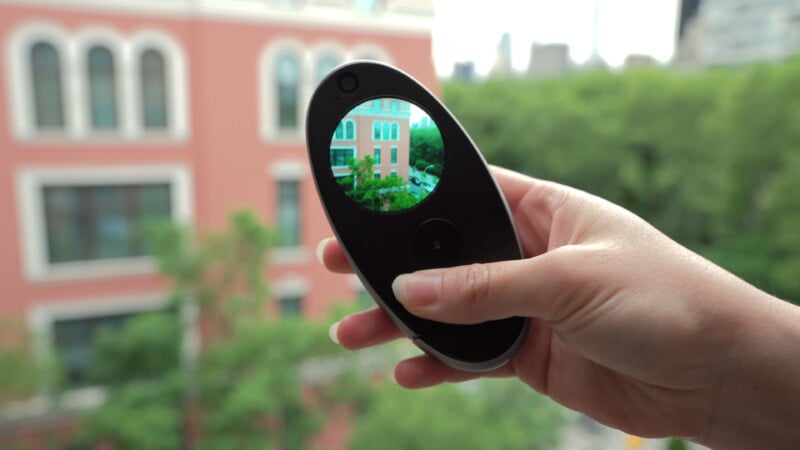 A hand holds a black digital viewfinder, displaying a sharp image of a pink building with white trim, while the background shows the same building out of focus with trees nearby.