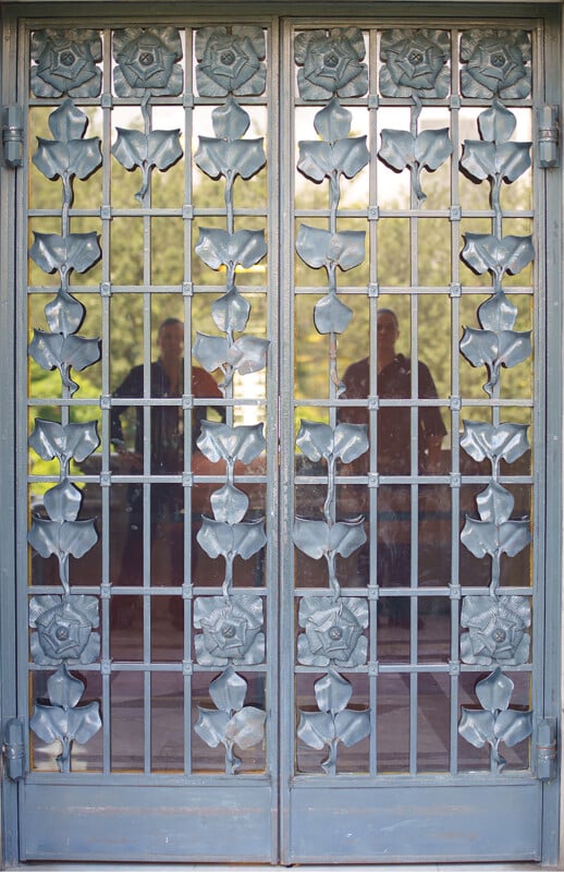 Metal double doors with a decorative floral pattern and glass panels. Two faint reflections of people are visible in the glass, and greenery can be seen outside through the door.