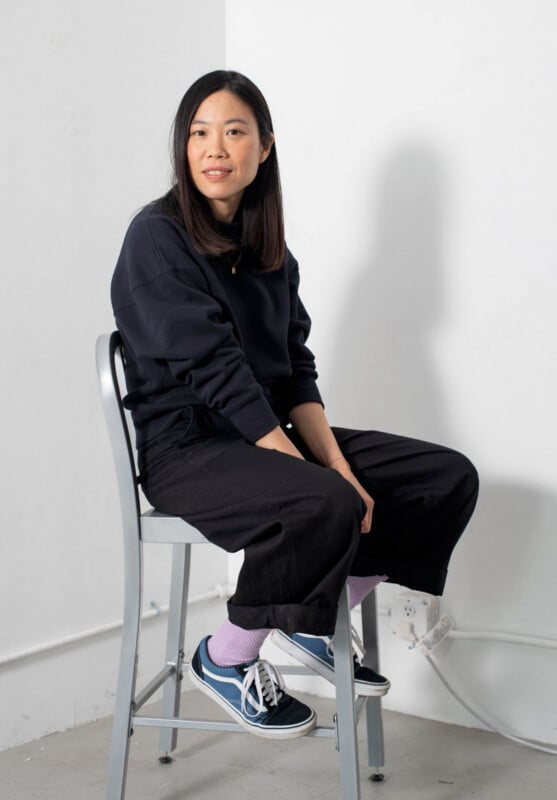 A woman with straight black hair sits sideways on a metal chair against a white wall. She wears a black sweater, black pants, purple socks, and blue sneakers, and looks calmly at the camera.