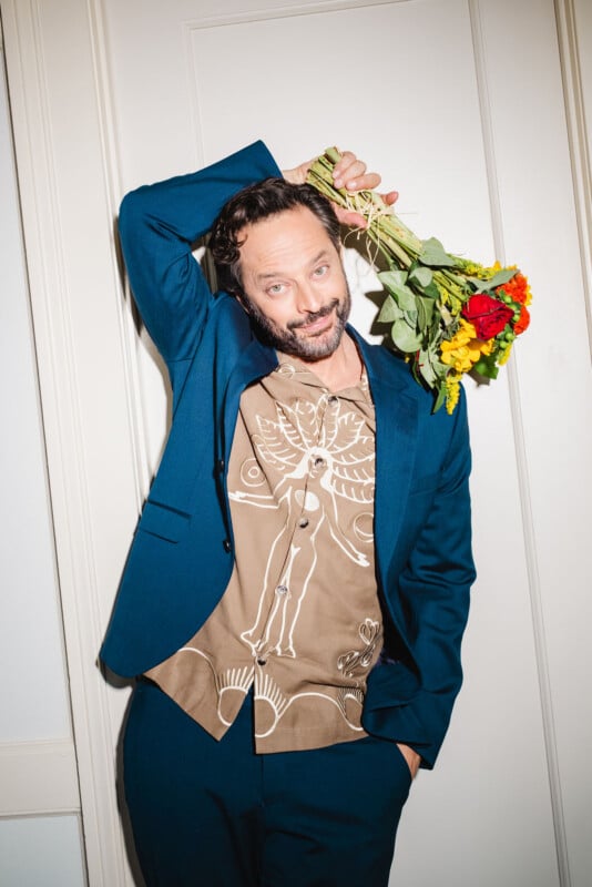 A man in a blue suit leans against a white wall, holding a bouquet of colorful flowers over his shoulder. He has a short beard, tousled hair, and wears a brown shirt with abstract line art.