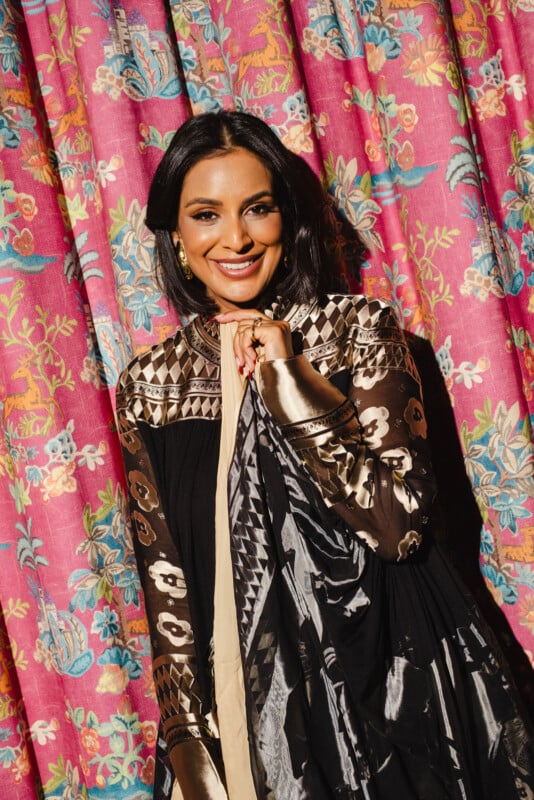 A woman in a black and gold patterned outfit stands smiling in front of vibrant pink floral curtains. She has dark hair, gold earrings, and holds part of her scarf near her face.