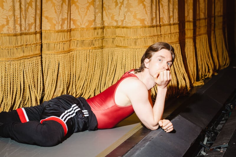 A person in a red tank top and black athletic pants lies on their side backstage, near gold curtains, resting their head on one hand and looking ahead thoughtfully.