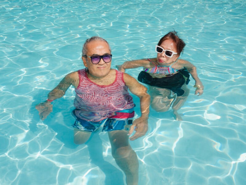 Two older adults wearing sunglasses and swimsuits relax together in a clear blue swimming pool, enjoying a sunny day. They are partially submerged in the water and appear calm and content.