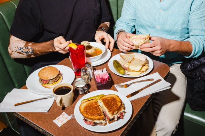 Two people sitting at a table with diner food: burgers, sandwiches, a pickle, coffee, a drink with lemon, soup, and packets of sugar. Both are mid-meal, with one person in black and the other in a light blue shirt.