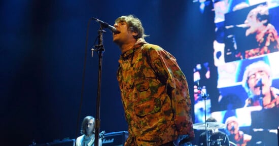 A singer in a colorful camouflage jacket leans into a microphone on stage. Behind him, a large screen displays multiple images of his face. Musicians and amps are visible in the background.