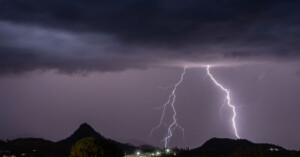 Several bolts of lightning illuminate a dark, cloudy night sky above distant hills and a small town, with silhouettes of trees visible in the foreground.