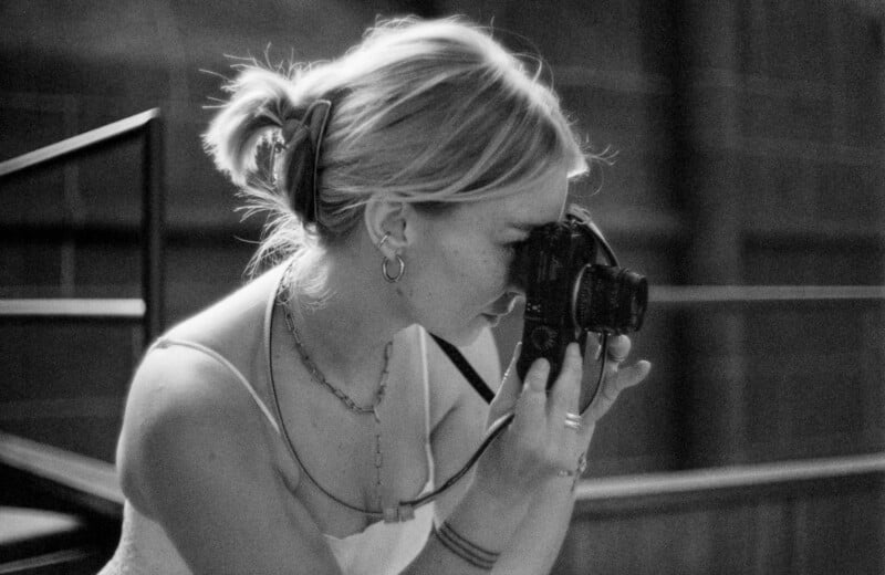 A woman with light hair in a ponytail holds a camera up to her face, appearing to take a photo. She wears hoop earrings, layered necklaces, and a sleeveless top. The image is black and white.