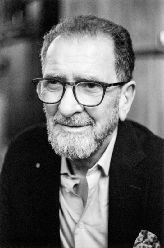 A grayscale photo of an older man with glasses, a beard, and short hair, wearing a suit jacket over an open-collared shirt, smiling slightly while looking off camera.