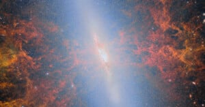 A bright spiral galaxy is seen edge-on, with vivid red and orange clouds of gas and dust radiating outward, surrounded by stars and cosmic dust against a dark space background.