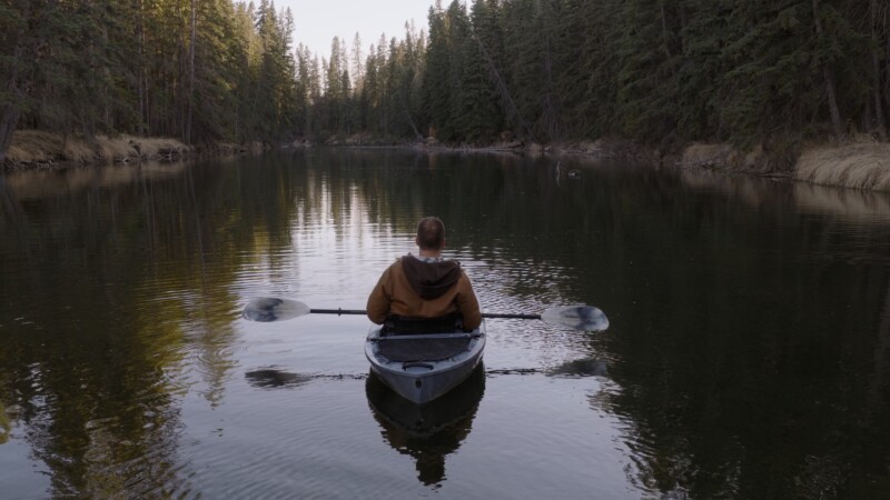A person paddles a kayak on a calm, tree-lined river. Tall evergreen trees flank both sides, reflecting in the water, and the scene feels peaceful and serene.