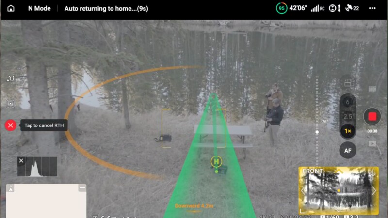 A drone’s return-to-home interface overlays a live aerial view of a lakeside scene with trees, a picnic table, and two people standing nearby. On-screen controls and a green path guide are visible.