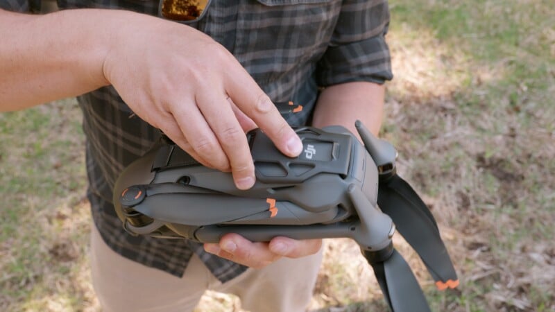 A person wearing a plaid shirt holds a folded drone with one hand and presses a button on top with the other hand, standing outdoors on grass.