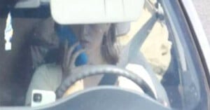 A person is driving a car while holding a phone to their ear, partially obscured by the rearview mirror and steering wheel. The person appears distracted while operating the vehicle.