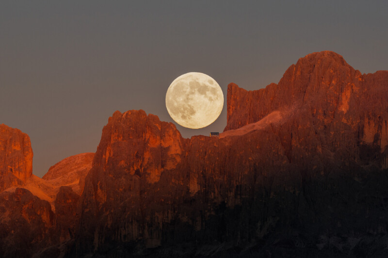 A large, full moon rises over rugged, reddish-orange mountain peaks at dusk, contrasting against the darkening sky. A small structure is silhouetted on the ridge below the moon.