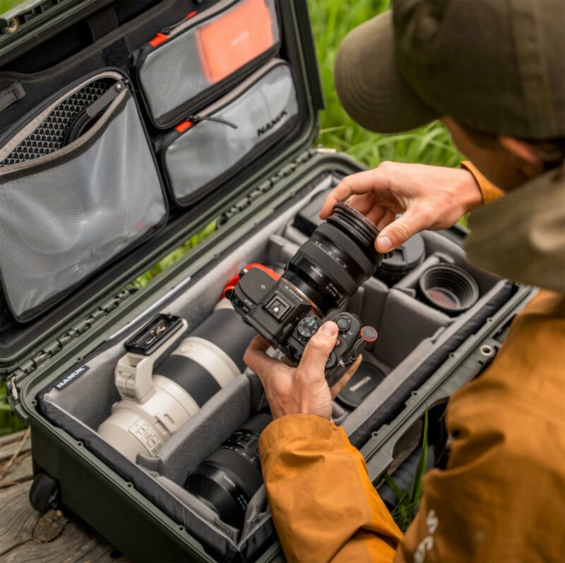 A person in an orange jacket places a lens on a camera, sitting next to an open padded camera case filled with camera gear and accessories on a grassy outdoor surface.