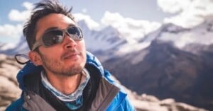 A man wearing sunglasses and a blue jacket looks up, with snow-covered mountains and a partly cloudy sky in the background.