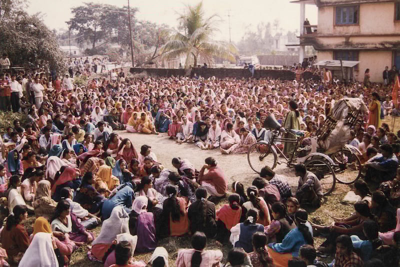 A large crowd of people, mostly women in colorful clothing, sit outdoors in a circle while a person on a tricycle appears to speak or perform in the center, with a building and palm trees in the background.