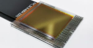 Close-up of a transparent, rectangular microchip or sensor with a grid pattern and circuitry, connected to a flat black ribbon cable at the top left.