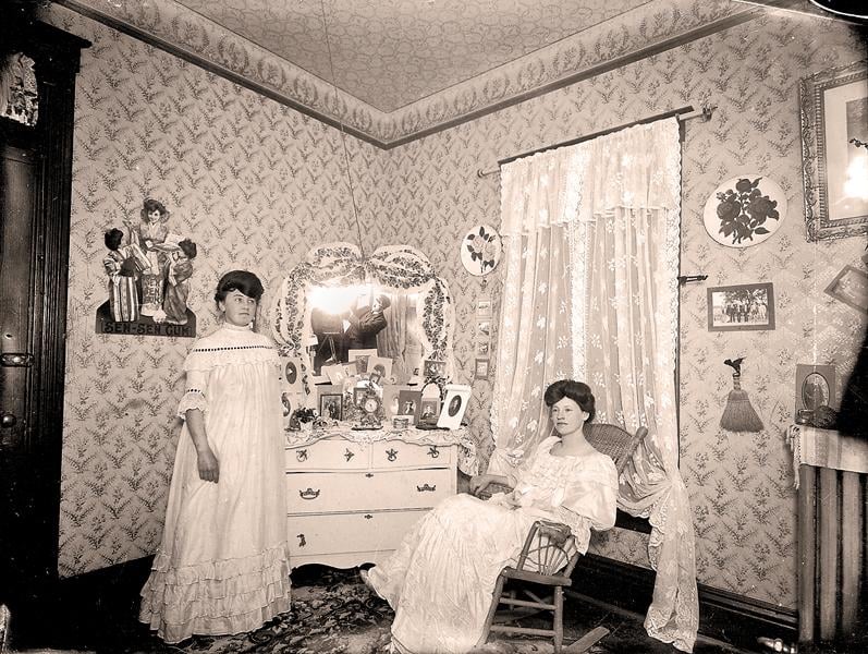 Two women in long white dresses pose in an ornate, old-fashioned bedroom with patterned wallpaper, a lace curtain, decorated vanity, framed photos, and various objects on display. One stands, the other sits in a rocking chair.