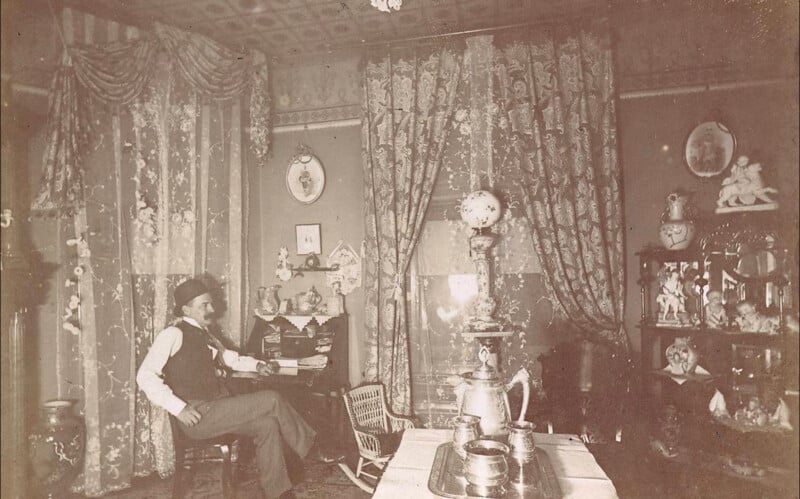 A sepia-toned photo shows a man in a hat and vest sitting in an ornate, Victorian-era living room with lace curtains, patterned wallpaper, and shelves filled with decorative items and porcelain figures.