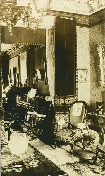 A sepia-toned photo of an ornate Victorian parlor featuring antique chairs, framed art on patterned walls, a piano, elaborate molding, and decorative rugs and curtains. The scene exudes vintage elegance and historic charm.