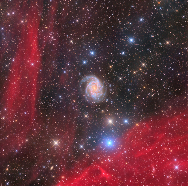 A colorful spiral galaxy surrounded by countless stars is set against a backdrop of red glowing nebulae and deep space. Bright blue and white stars are scattered throughout the image.