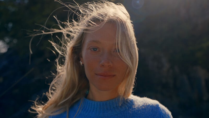 A woman with long blonde hair stands outdoors, wearing a blue sweater. Sunlight softly illuminates her face and hair, and the background is blurred with natural scenery.