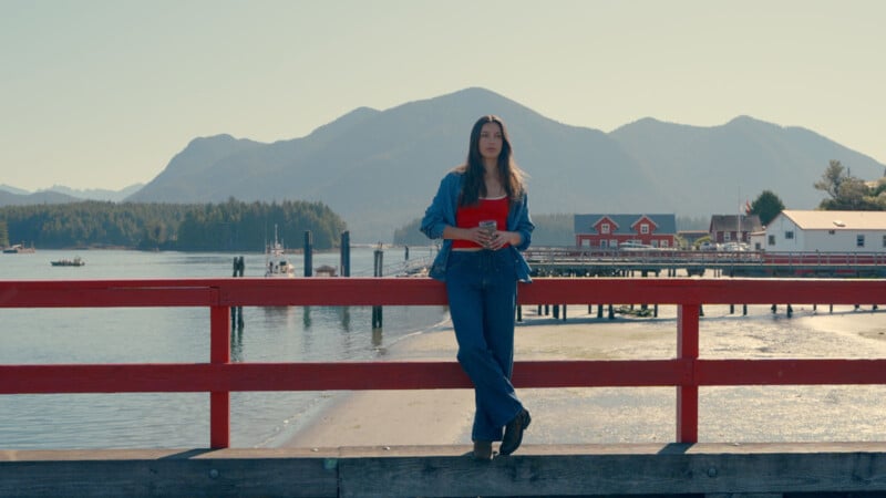 A woman in blue jeans and a denim jacket stands holding a red notebook on a red railing by the water, with mountains and houses in the background on a sunny day.