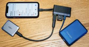 A smartphone connected via adapter hub to various devices including an external hard drive, an SSD, and an SD card, all placed on a wooden surface. The phone screen shows a file explorer app.
