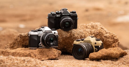 Three OM System OM-5 cameras in black, silver, and gold are displayed on rugged brown rocks in an outdoor setting with a blurred sandy background.