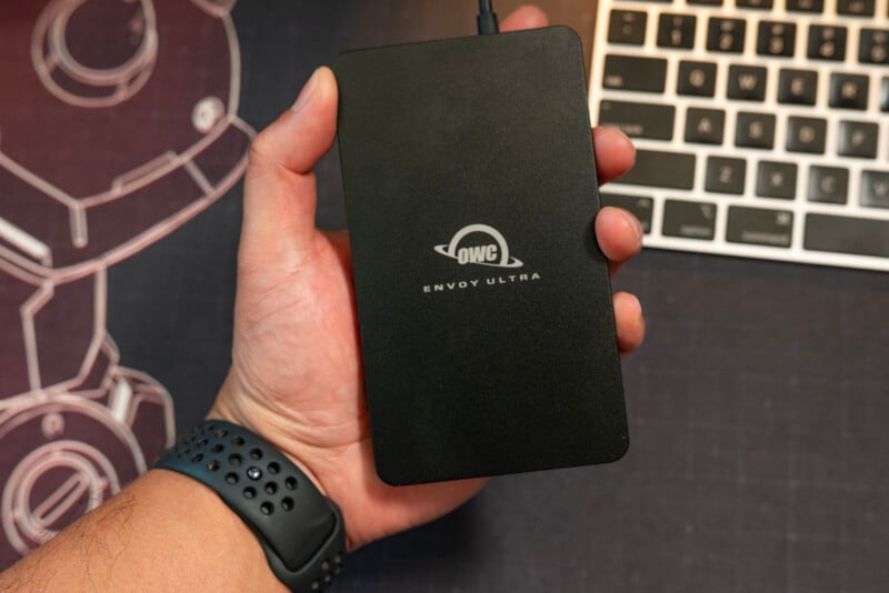 A person wearing a black smartwatch holds a black OWC Envoy Ultra external hard drive. Part of a laptop keyboard and a technical drawing are visible in the background.