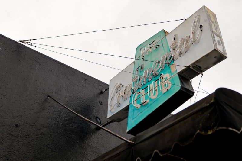 A vintage turquoise and white neon sign reading "The Continental Club" is mounted on the side of a dark building against a cloudy sky. The sign appears weathered and is supported by metal rods.