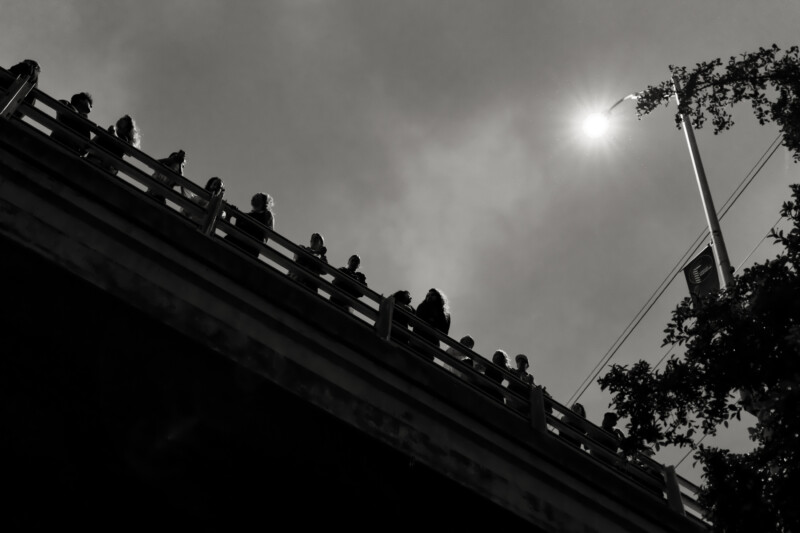 Silhouetted people stand along the edge of a bridge under a cloudy sky, with the sun shining brightly near a streetlight and tree branches visible on the right.