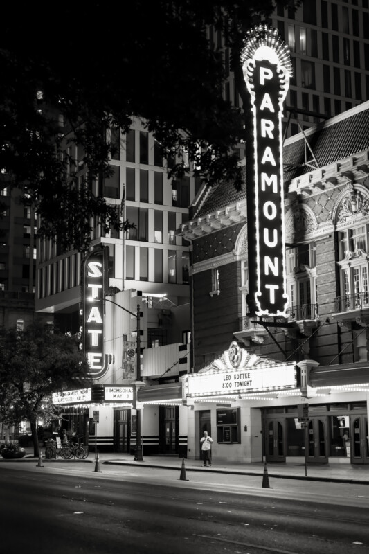 Black-and-white photo of a nighttime city street with the illuminated Paramount and State theater marquees. The Paramount sign is vertical and prominent, and the vintage theater fronts are lit, with a few people visible on the sidewalk.
