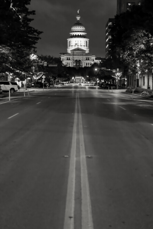 A black and white photo of an empty street at night, leading directly to a domed government building illuminated in the background, framed by trees and city buildings.