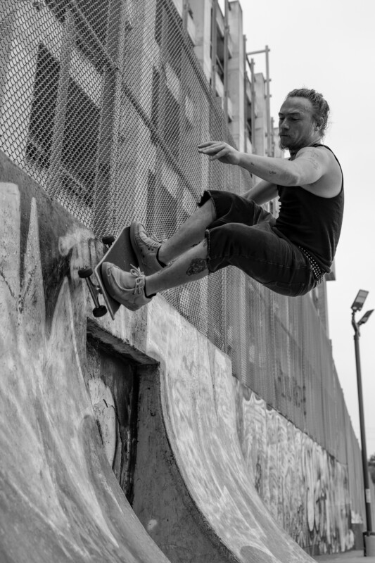 A skateboarder with tattoos and a beard rides up the curved wall of an urban skate park, captured mid-air beside a chain-link fence and graffiti-covered concrete in black and white.