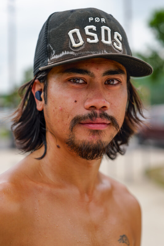 A young man with long dark hair, a mustache, and beard wears a black "OSOS" cap and a wireless earbud. He is shirtless, looking directly at the camera, with water droplets visible on his skin.