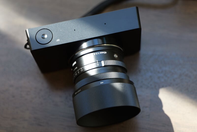 A modern black digital camera with a large lens sits on a wooden surface. Sunlight casts soft shadows, highlighting the camera's sleek design and textured focus ring. The lens is extended outward, suggesting readiness for use.