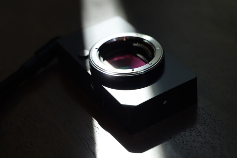 A close-up of a black, rectangular camera body with a circular lens mount. It is partially illuminated by a beam of light, casting a shadow on a dark surface.