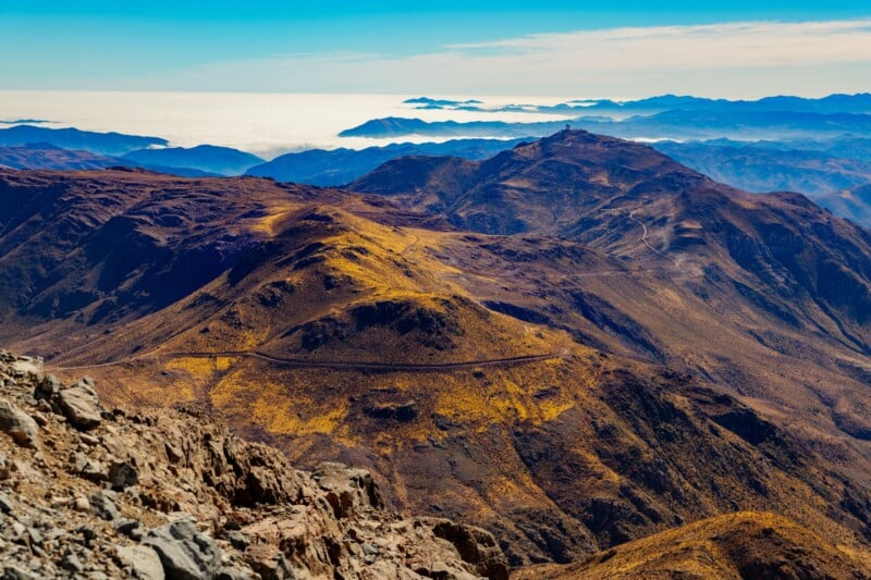 A panoramic view of rugged, sunlit mountains with a winding road, golden grassy slopes, and distant mountain ranges under a blue sky above a layer of clouds near the horizon.