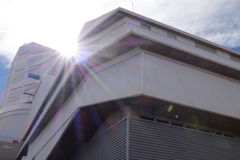 Sunlight shines through the upper corner of a modern, angular industrial building with metal siding, creating lens flares across the image under a partly cloudy sky.
