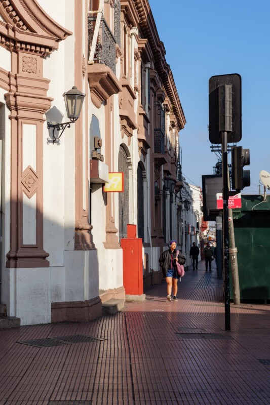 A person walks on a sunlit city sidewalk beside ornate, historic buildings with balconies and decorative columns. Street signs, lampposts, and a green dumpster are visible along the path.