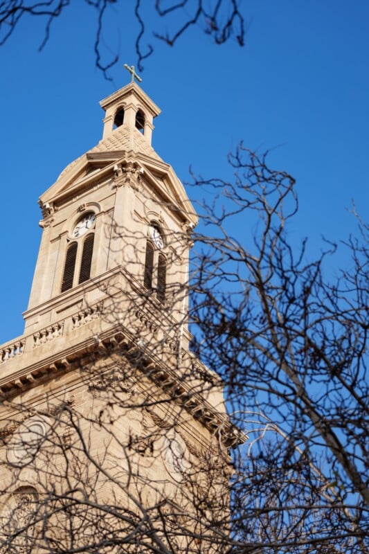 A tan stone church tower with arched windows and a small cross on top is seen through leafless tree branches against a clear blue sky.