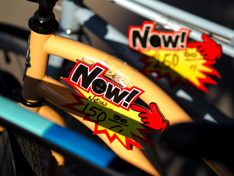 Close-up of a yellow bicycle frame with a red and black "Now!" sale sign attached, showing a discounted price and a red pointing hand, with another similar sign visible in the background.