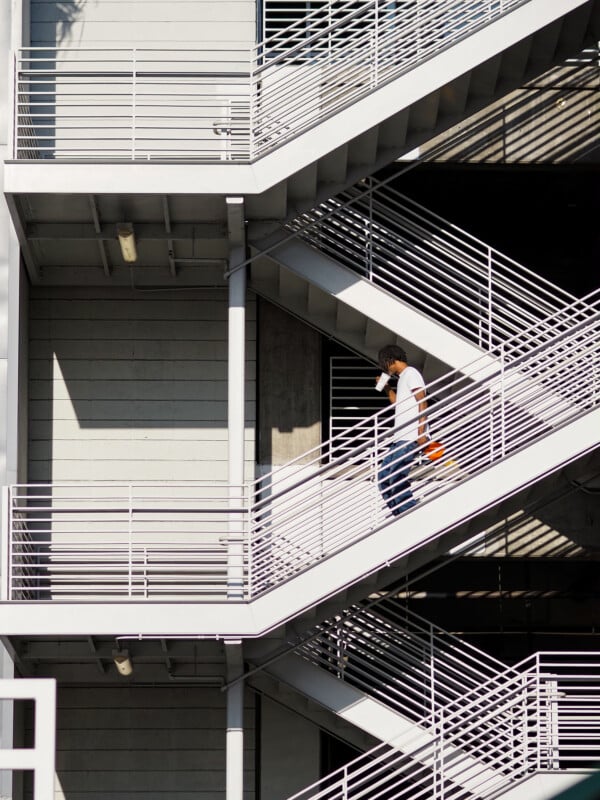 A person walks down an outdoor, zigzagging metal staircase attached to a building, holding an orange object and shielding their face from the sun. Shadows create geometric patterns on the gray walls.